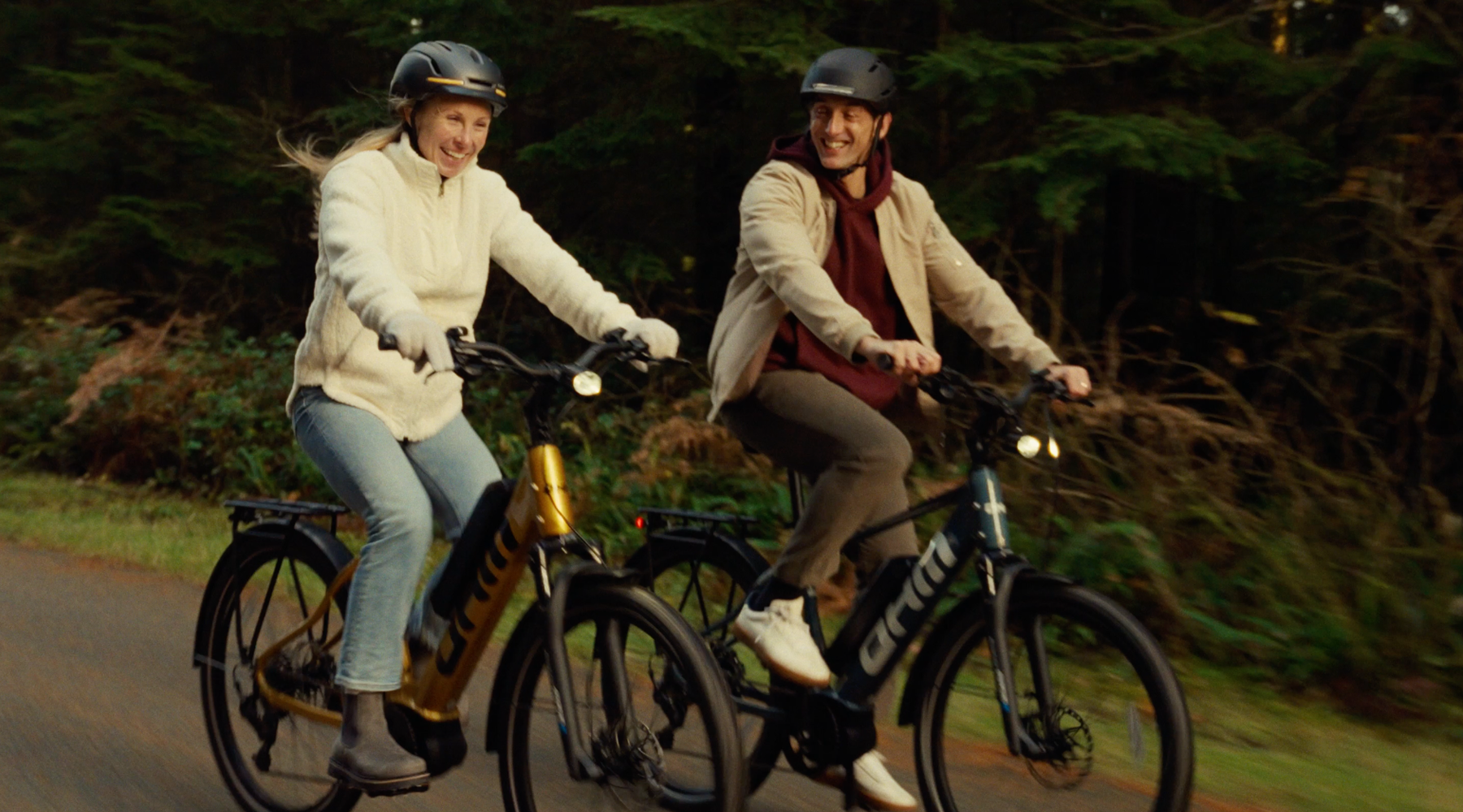 Two riders cycling side by side on OHM Discover e-bikes through a sunlit Pacific Northwest forest trail